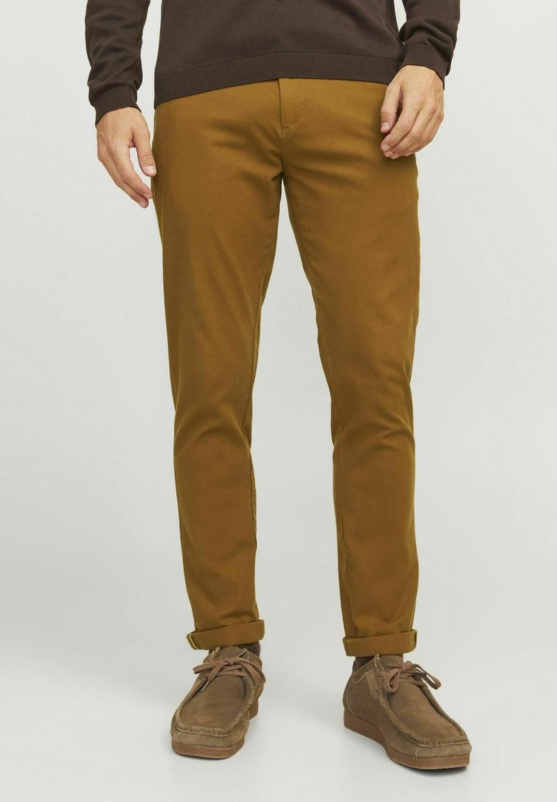 Pantalons en coton marron avec une coupe slim, présentant des poignets roulés. Associés à des chaussures en suède marron. Texture lisse et design décontracté.