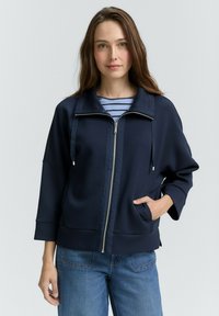 Mujer con cabello castaño largo que lleva una chaqueta azul marino con cremallera sobre una camiseta de rayas azules y vaqueros azules, de pie con una mano en su bolsillo.