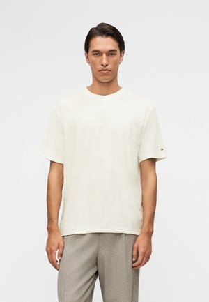 Jeune homme aux cheveux courts et foncés portant une chemise blanche à manches courtes ample et un pantalon gris clair, debout devant un fond uni clair.