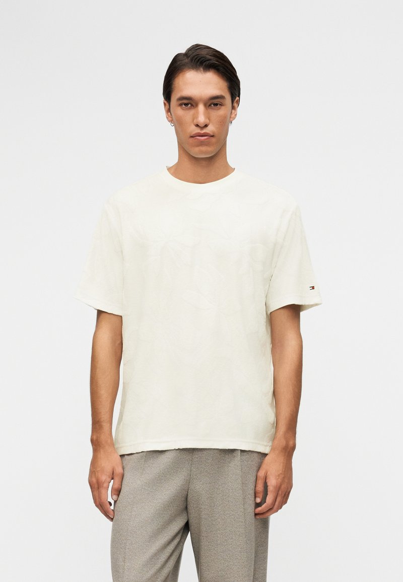 Νεαρός άνδρας με κοντά σκούρα μαλλιά φοράει ένα άνετο λευκό πουκάμισο με κοντά μανίκια και ανοιχτό γκρι παντελόνι, στέκεται μπροστά σε ένα απλό φωτεινό φόντο.