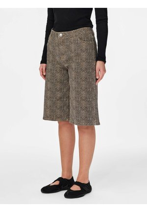 Persoon draagt knielange korte broek met luipaardprint, zwarte top met lange mouwen en zwarte riem schoenen, staand tegen een effen achtergrond.