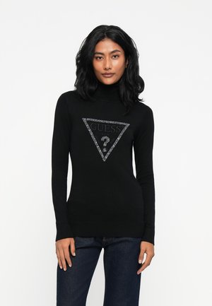 Donna con capelli neri e ricci indossa un maglione a collo alto nero con un logo GUESS glitterato argento e jeans blu scuro, in piedi contro uno sfondo bianco.