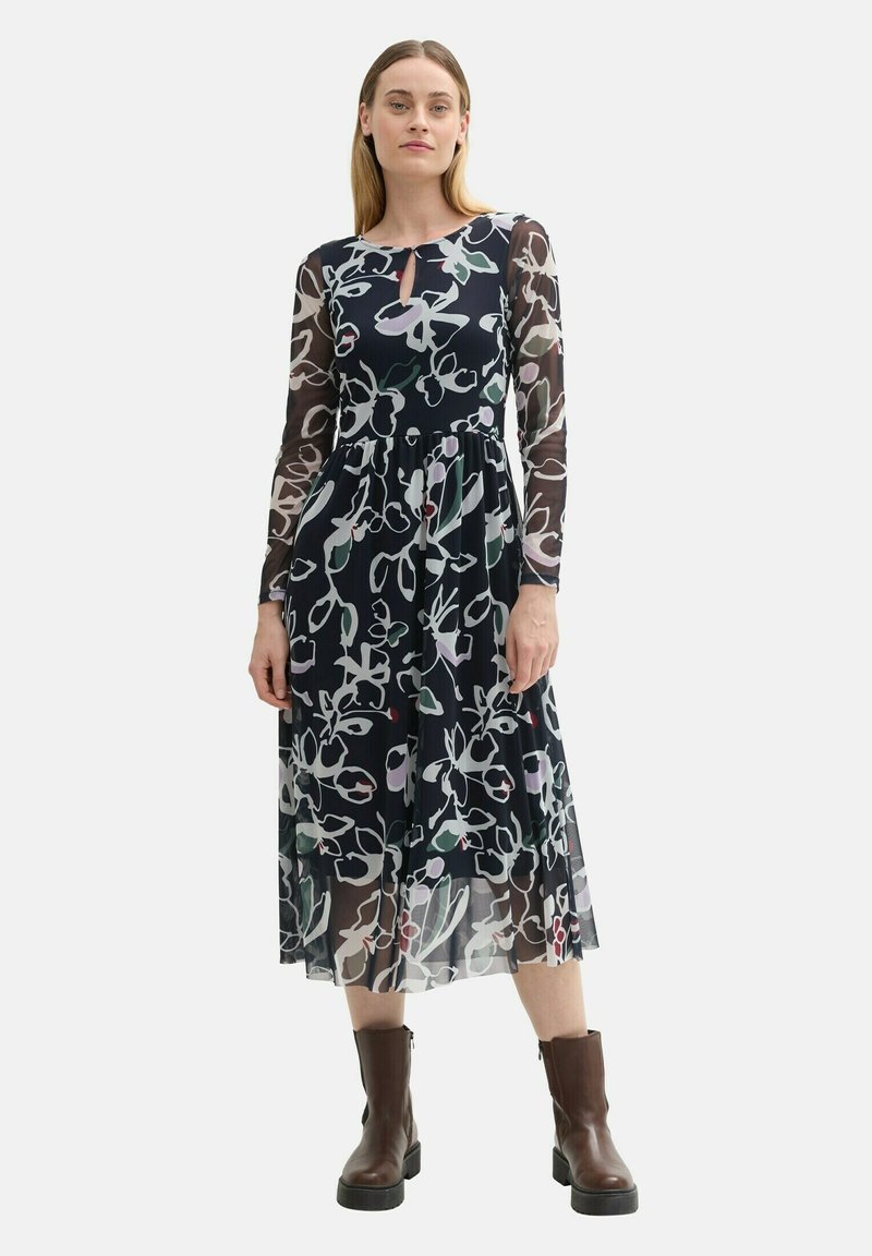 Blumengemustertes Kleid mit langen transparenten Ärmel, einem marineblauen Hintergrund und einem Schlüsselloch-Ausschnitt. Kombiniert mit dunkelbraunen Knöchelstiefeln.