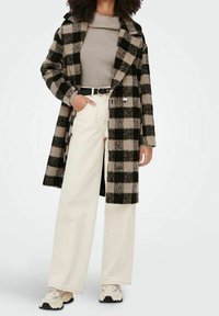 Manteau à carreaux noir et beige au design ajusté, accompagné d'un pull beige côtelé, d'un pantalon large crème et de baskets beiges.