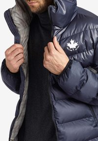 Uomo che indossa una giacca puffer blu navy con fodera a scacchi, che sistema la cerniera, mostrando il logo bianco della foglia d'acero e il testo "For a Few EST 2001".