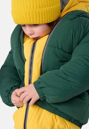 Giacca imbottita verde e gialla con chiusura a zip, con polsini elastici. Il modello sta chiudendo la giacca mentre indossa un berretto giallo.