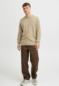 Beige knitted sweater with a round neckline, worn with loose brown pants and gray athletic shoes featuring a textured design.