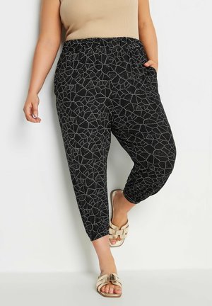 Pantalons noirs à taille élastique avec un motif géométrique blanc, poches latérales, chevilles effilées et texture douce, associés à des sandales neutres.