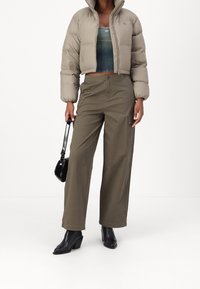 Giacca a vento beige, corta, con colletto alto; indossata sopra un top fitted blu-verde. Pantaloni a gamba larga oliva con tasche, stivaletti neri con tacco.