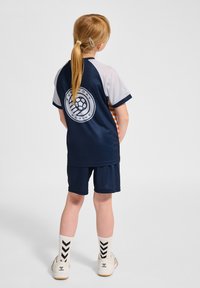 Mörkblå sporttröja med grå ärmar, med vitt logotyp och randig detalj. Kombinerad med matchande shorts och randiga strumpor.