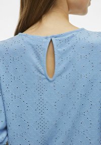 Ljusblå blus med öppen nyckelhålsrygg, texturerad tyg med mönstrad broderi och korta ärmar.