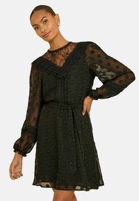 Černé průhledné šaty s texturou puntíků, krajkovým lemem a dlouhými rukávy. Vybavené úzkým pasem s vázacími pásky a volánkovými detaily na výstřihu.