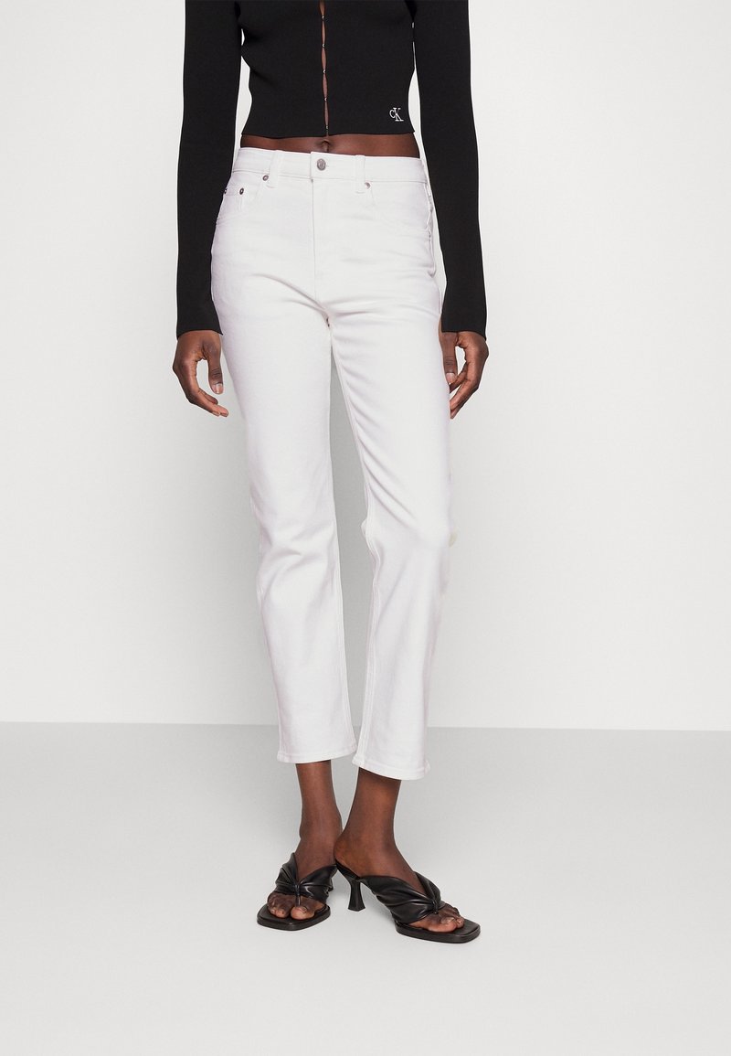 Personne vêtue d'un top noir court à manches longues, d'un jean blanc taille haute à jambes droites, et de sandales noires à talons ouverts, debout devant un fond uni.