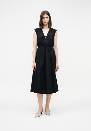 Žena stojaca v čiernych midi šatách bez rukávov s predným viazaním a svetlosivými nízkymi lodičkami so špicatou špičkou na bielom pozadí.