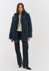 Žena v modrom kožuchu so strapcami, krémovom pletenom svetri, čiernych širokých nohaviciach a čiernych špicatých topánkach na bielom pozadí.