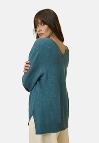 Teal ulltröja med V-ringning, långa ärmar och sidoflikar. Mjuk textur med ribbat nederkant. Modellen visar bakvyn.