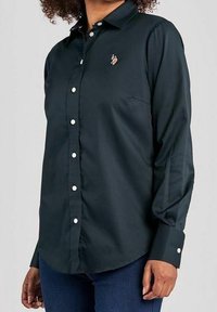 Mörkgrön skjorta med knappar, tillverkad av ett slätt tyg, med klassisk krage och långa ärmar. Har vita knappar och en liten logotyp på bröstet.