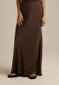 Brązowa, zwiewna spódnica maxi z gładkiego materiału o lekko połyskującej powierzchni, z bocznym rozcięciem, noszona z czarnymi klapkami.