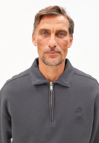 Middelmatige man met lichte stoppels en blauwe ogen, die een donkergrijze sweatshirt met rits en kraag draagt tegen een witte achtergrond.