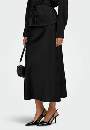 Femme portant une longue jupe noire, un chemisier noir, des talons slingback noirs, tenant un petit sac à main noir sur un fond uni.