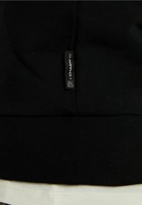 Svart sweatshirt med ribbad textur, med en sydd fåll och en synlig etikett som läser "CHASIN' 92." Positionerad ovanför ett vitt tyg.