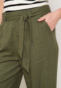 Olive green, linen blend trousers with a wide waistband and a tied belt. Features side pockets and a relaxed fit. Striped shirt visible above.