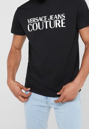 Man draagt een zwart t-shirt met de witte tekst "Versace Jeans Couture", gecombineerd met lichtblauwe spijkerbroek, en past de zoom van zijn overhemd aan.