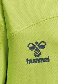 Limegrönt atletiskt tyg med en strukturerad yta, med en mörkblå logotyp av en bi och varumärket "hummel" tydligt presenterat.