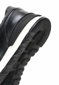 Zapatilla de cuero negra con una parte superior texturizada, talón moldeado y una entresuela blanca en contraste, junto con una suela de goma con patrón.