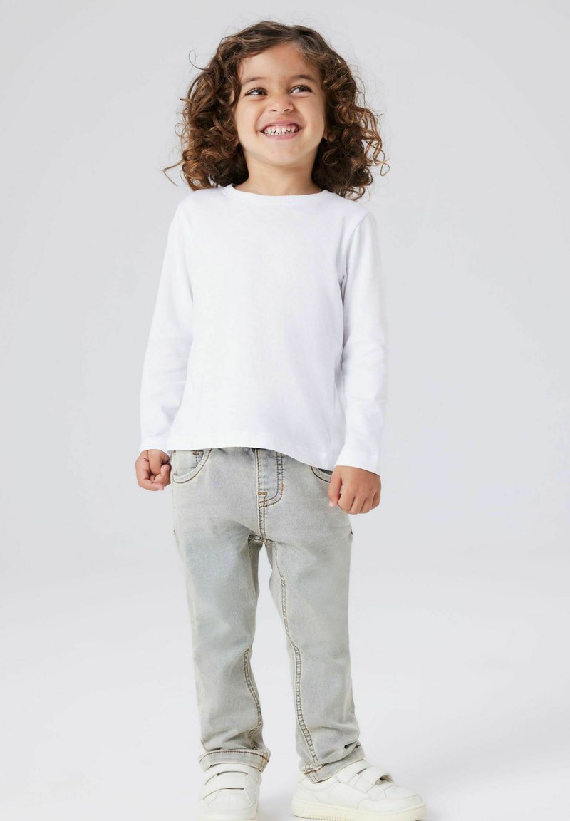 Bela majica z dolgimi rokavi, svetlo modri jeans hlače in beli supergi. Otrok stoji nasmejan z kodrastimi lasmi, roke ob telesu. Preprost dizajn.