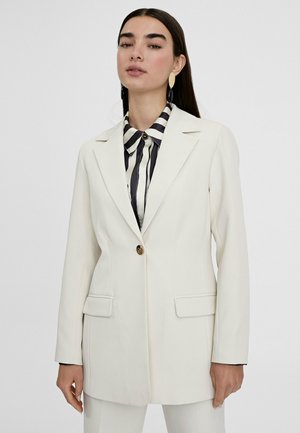 Witte getailleerde blazer met een enkele gouden knoop, notch-revers en twee voorzakken, gecombineerd met een zwart-wit gestreept shirt eronder.