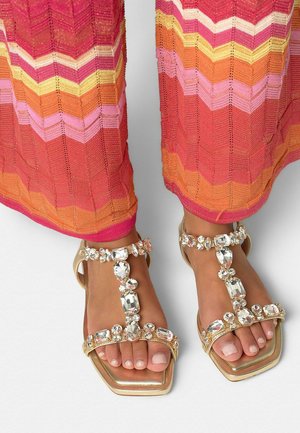 Pies con sandalias doradas adornadas con grandes piedras preciosas transparentes, combinadas con pantalones a rayas de colores en tonos rosa, naranja y amarillo.
