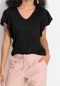 Woman wearing a black V-neck flutter sleeve top tucked into high-waisted light pink pants with a tied waist, hand in pocket, on white background.