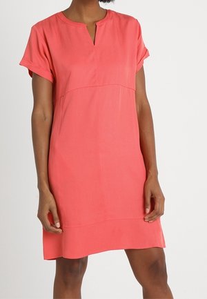 Femme portant une robe corail à manches courtes avec un petit col en V et des poignets retroussés, debout devant un fond clair uni.