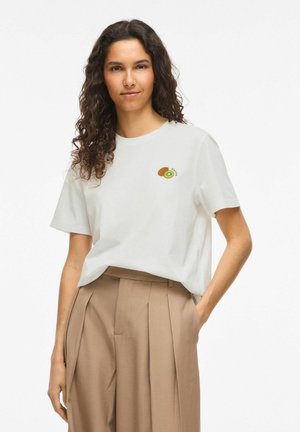 Femme aux longs cheveux bouclés portant un t-shirt blanc avec un petit motif kiwi et un pantalon beige plissé taille haute, debout avec une main dans la poche.