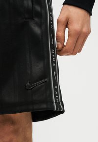 Δεξί χέρι δίπλα σε μαύρο σορτς Nike με κάθετη λωρίδα λογότυπου και κεντημένο σήμα Swoosh στο κάτω μέρος.