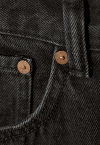 Detailní záběr na černou denimovou látku zobrazující okraj kapsy, poutko na opasek, švy a měděné nýty na džínách.