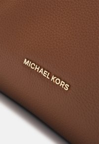 Brązowa skórzana faktura z wyraźnie wyeksponowanym złotym logiem "MICHAEL KORS". Subtelny wzór ziarna podkreśla walory wizualne.
