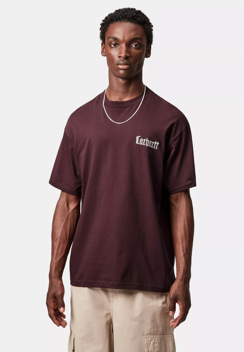 T-shirt in cotone bordeaux con maniche corte, con logo bianco "Carhartt" sul lato sinistro del petto. Il modello indossa una collana d'argento.