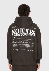Sudadera marrón oscuro con un gran gráfico de texto blanco en la espalda, que presenta varias frases motivacionales. Tejido suave, corte relajado, mangas largas.