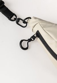 Hellbeige Tasche mit einem schwarzen Reißverschluss, ausgestattet mit einem schwarzen Tragegurt und metallischen Beschlägen. Das Design umfasst eine geschwungene Form und eine strukturierte Stoffoberfläche.