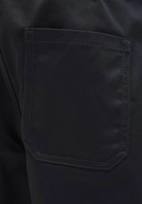 Gros plan d’un pantalon en tissu noir montrant une poche arrière cousue sur le côté droit.