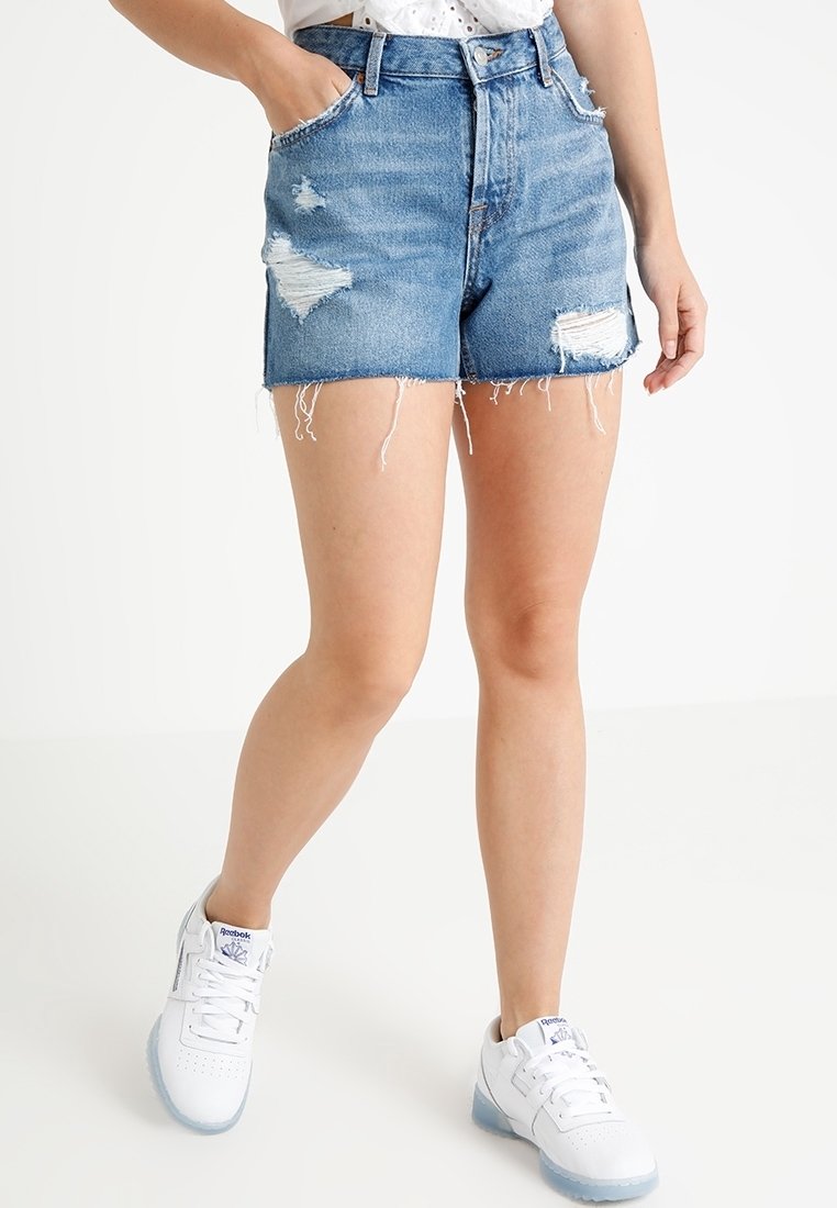 Džíny šortky s vysokým pasem, světle modré barvy, s opotřebovanými detaily, roztrhaným lemem a přiléhavým střihem. Nosené s bílými teniskami.