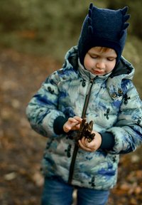 Bērns rakstainā pūkainā jaciņā, turot nūjas, valk�ājot tumši zilu adītu cepuri ar teksturētām detaļām. Āra vide ar izplūdušu fonu.