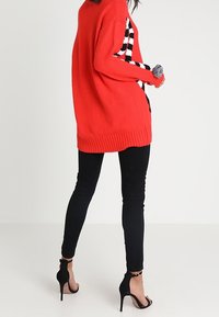 Cardigan rosso lavorato a maglia con una vestibilità morbida, caratterizzato da un pannello laterale a quadretti bianco e nero. Jeans skinny neri e sandali con tacco alto completano il look.