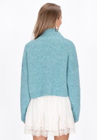 Zkrátený sveter v svetlo teal farbe, s rebrovanou textúrou, vysokým golierom a spadnutými ramenami, ktorý je kombinovaný s bielymi šatami s čipkovým lemovaním.