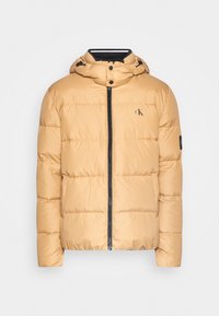 Puffjakke i lys tan med lynlås foran, aftagelig hætte og syede sektioner. Har et lille logo på brystet og sorte detaljer.