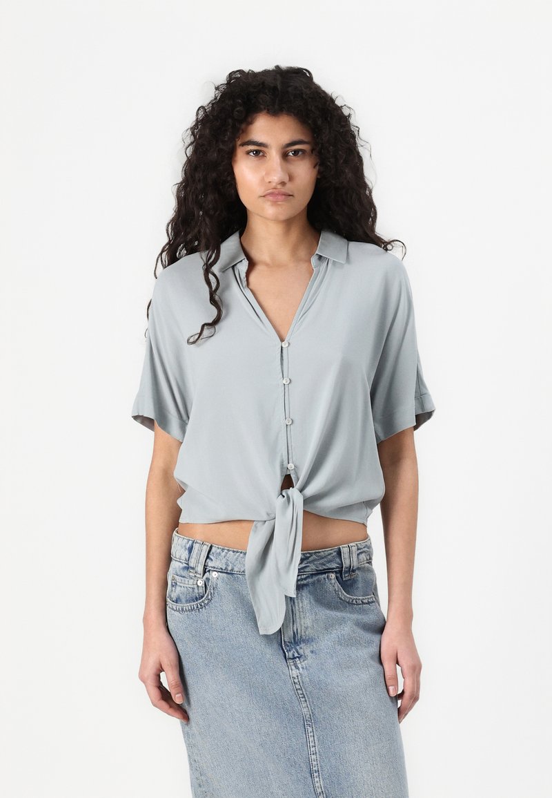 Light grey cropped blouse with a collar, short sleeves, and a front tie detail, made from a smooth fabric. Paired with a denim skirt.