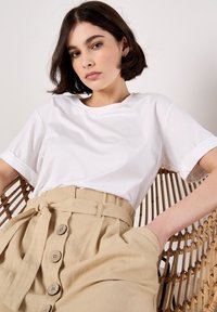 T-shirt en coton blanc avec des manches retroussées, associé à une jupe en lin beige avec une ceinture nouée et cinq boutons sur le devant. Fond texturé en assise.