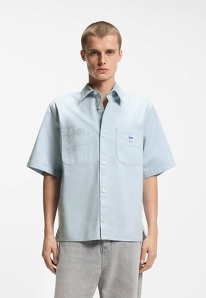 Ung mand med kort hår iført en lyseblå kortærmet skjorte med to forlommer og lysegrå bukser.